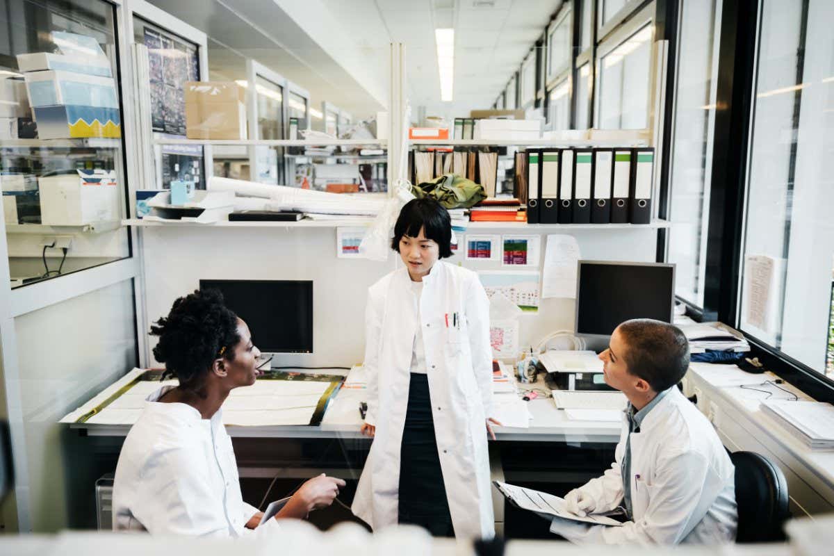 An Asian, a black and a white female scientist are in a laboratory wearing typical white lab clothes. They are discussing their research surrounded by files and computers. One of them is holding a clipboard.