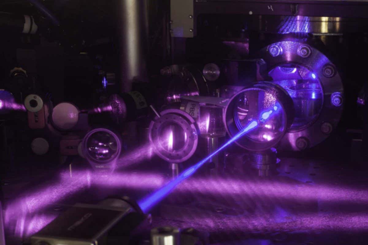 JILA?s three-dimensional (3-D) quantum gas atomic clock consists of a grid of light formed by three pairs of laser beams. A stack of two tables is used to configure optical components around a vacuum chamber. Shown here is the upper table, where lenses and other optics are mounted. A blue laser beam excites a cube-shaped cloud of strontium atoms located behind the round window in the middle of the table. Strontium atoms fluorescence strongly when excited with blue light.