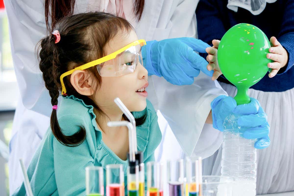 Portrait closeup shot of Asian little curious elementary girl with bunches pigtail hairstyle wears safety goggles looking at water bottle blowing green rubber balloon experiment teach by teacher.; Shutterstock ID 1985510477; purchase_order: 30 July 2022 issue; job: Photo; client: NS; other: