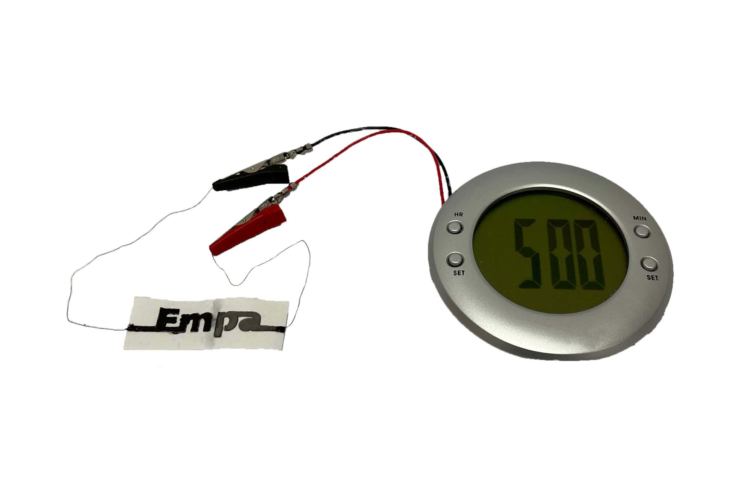 Photograph of a stencil-printed two-cell paper battery with a design that spells the name of the authors' research institution (Empa). The battery is running a liquid crystal display alarm clock. Credit: Alexandre Poulin.