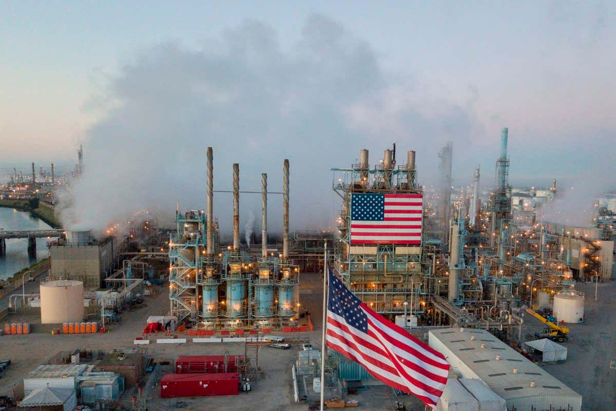 Steam rises from an oil refinement facility. Two American flags are displayed in an industrial setting with many pipes, tanks, and buildings.