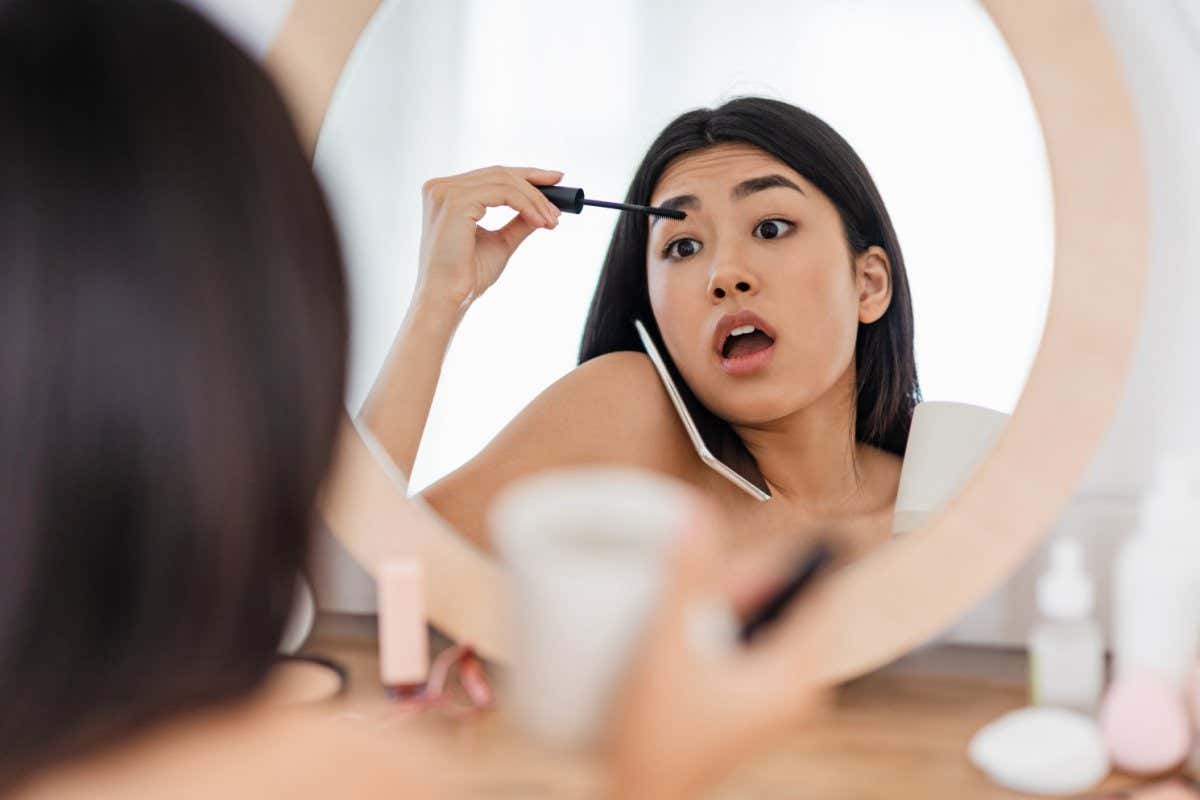Impressed young asian woman talking on phone with girlfriend while putting mascara in front of mirror at home, sharing rumors; Shutterstock ID 1793035801; purchase_order: -; job: -; client: -; other: -