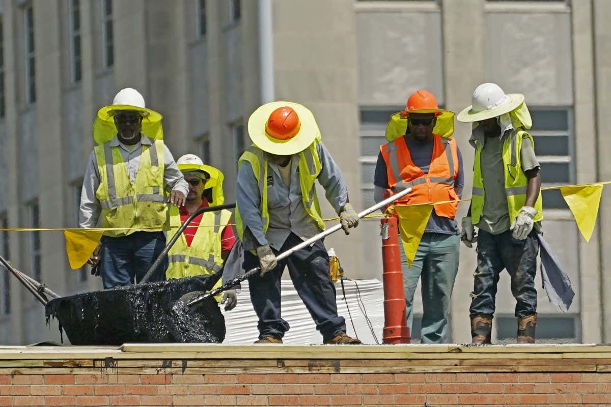 Five construction workers wearing yellow and orange vests and wide-brimmed helmets repair a roof, while one worker in the middle spreads hot tar on the roof's surface.