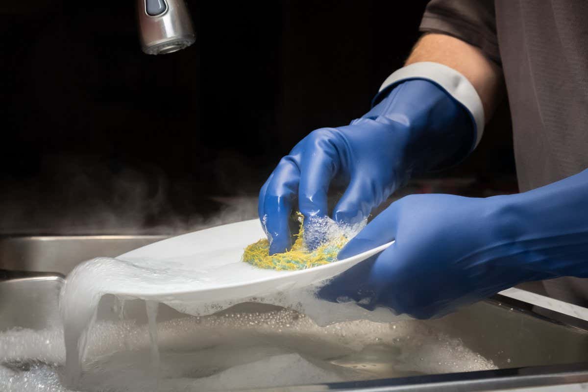Close up of a model's hands washing dishes in steaming hot water with dark blue rubber gloves with anit-drip cuffs
