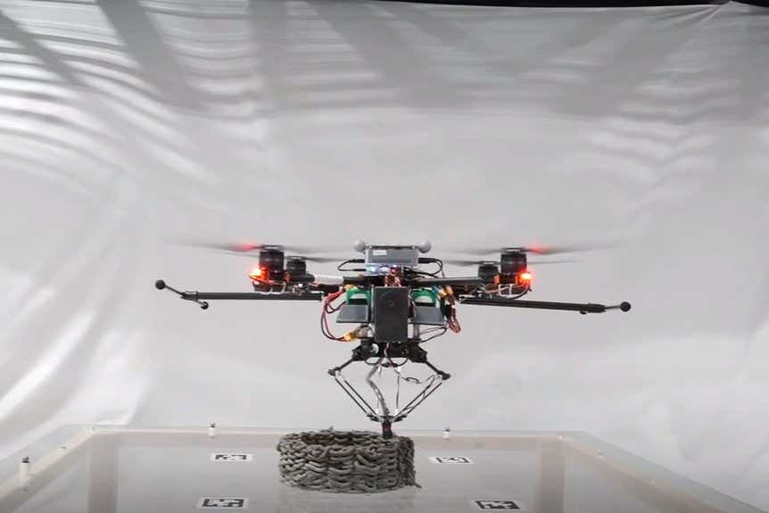 A specialized robotic drone sprays layers of cement to create a cylindrical structure in a clean laboratory setting with white background behind a staging area for the structure's construction