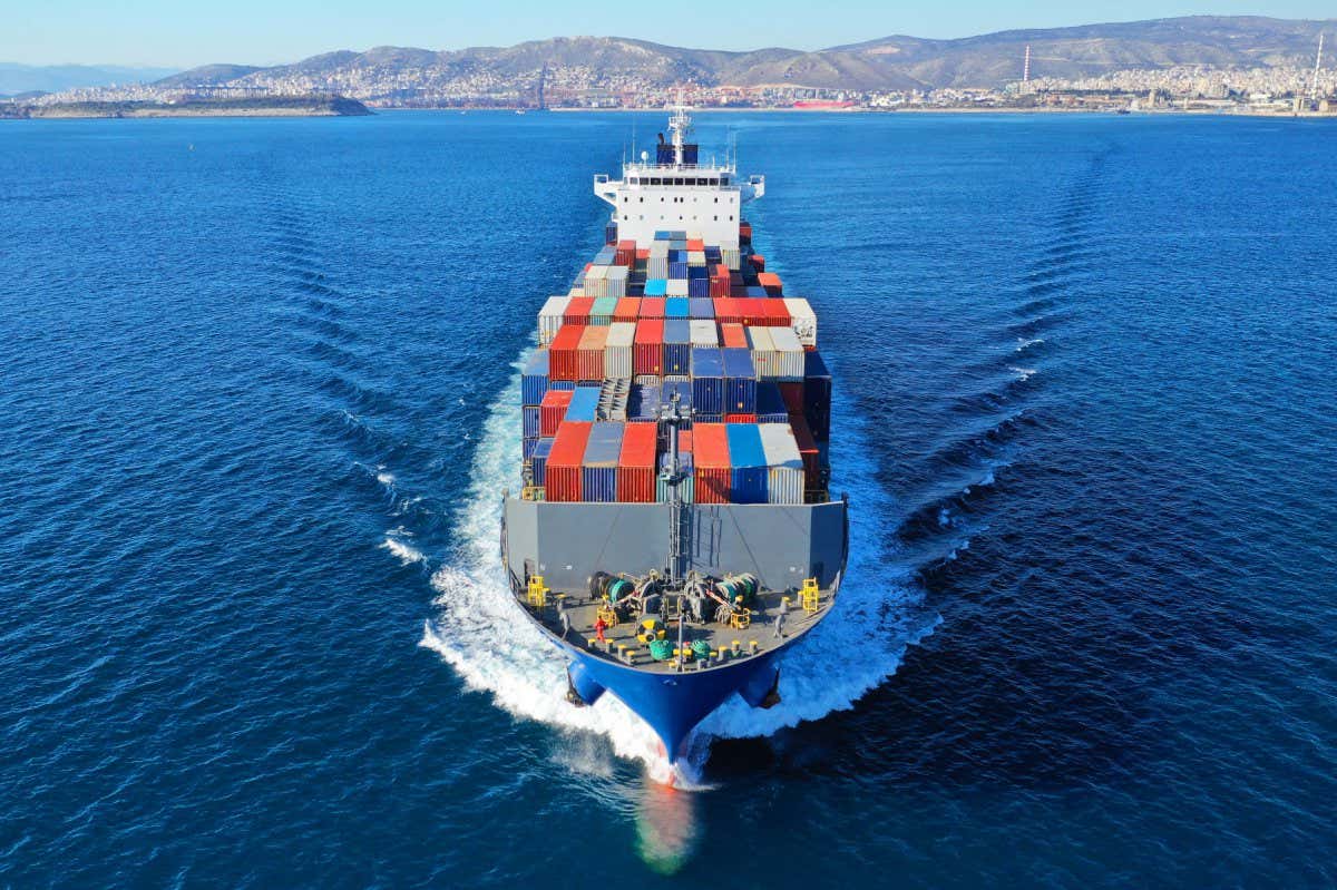 An above view of the front of a container ship moving through deep, blue ocean waters on a sunny day with dry hills in the background on shore. The ship is fully loaded with colourful, truck-sized cargo containers.