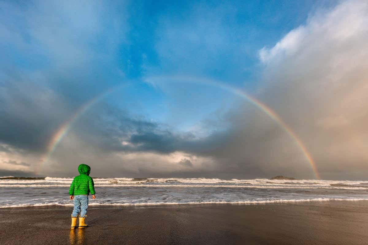 2GPFM5E Child in green jacket looking at rainbow over an ocean