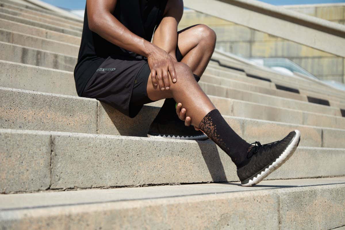 Sports injury concept. Cropped portrait of black male runner wearing black training outfit touching his leg in pain with clasped hands, having sprain or twitch in his knee after running exercises; Shutterstock ID 478297546; purchase_order: -; job: -; client: -; other: -