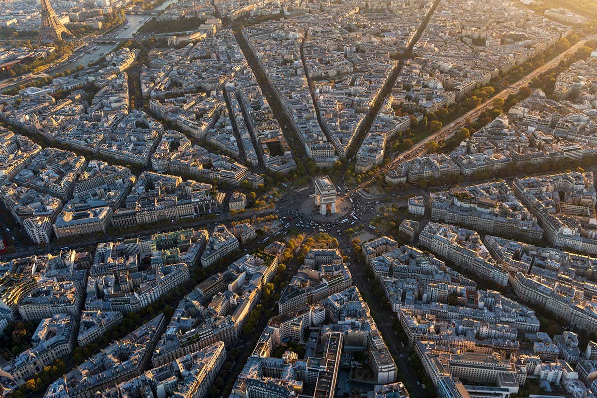 An aerial photo taken over the Arc de Triomphe in Paris, France at sunset with a wide view above the monument, with streets coming out like spokes from the monument at the center.