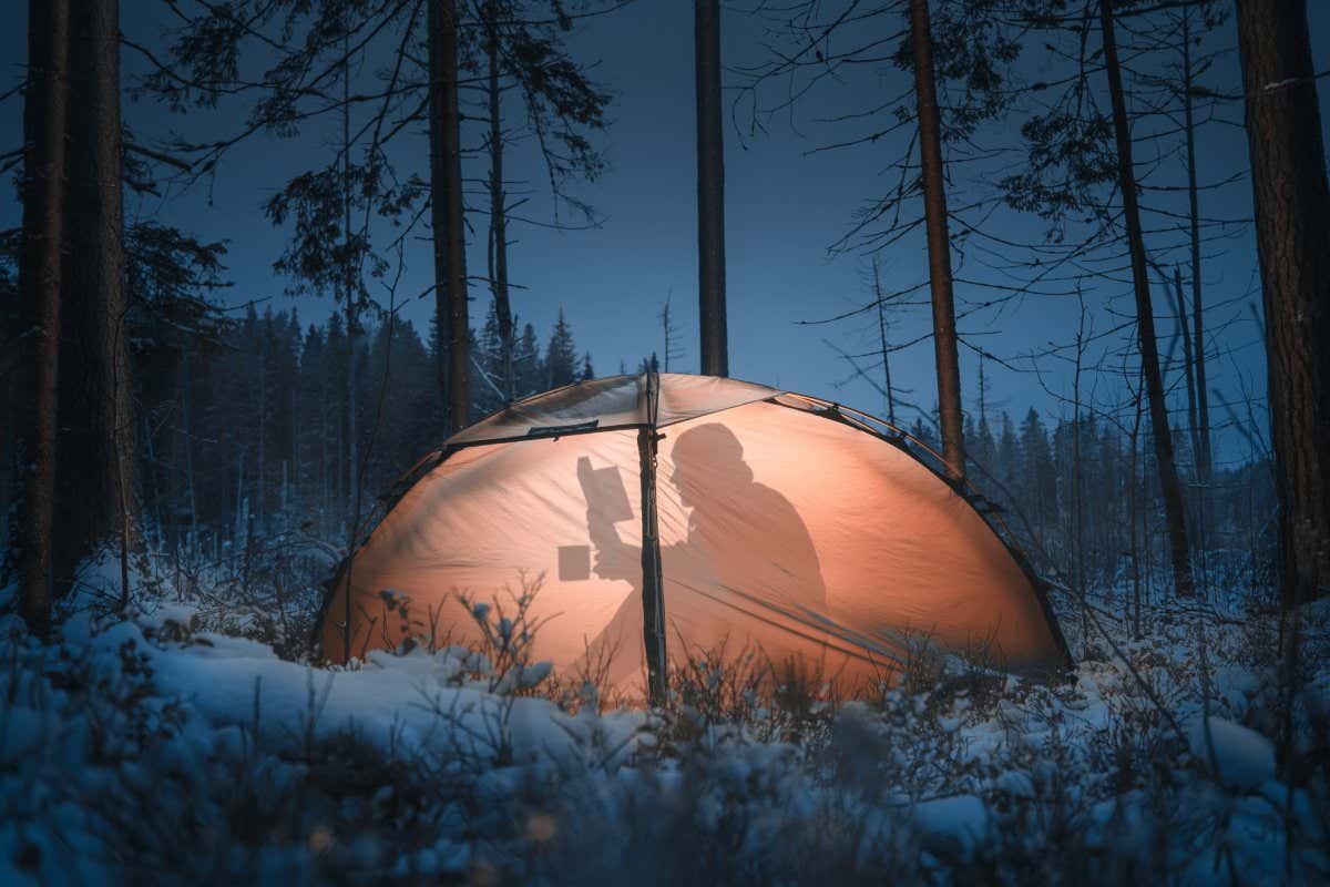 Silhouette of a man in a tent. He reads a book and drinks tea. Winter time. Pine forest.
