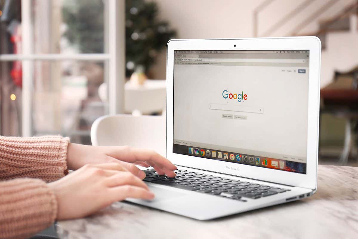 A laptop sits on a desk, opened to the homepage for the search engine Google. A person's hands enter from the left, with one hand's fingers placed on the laptop's keyboard.
