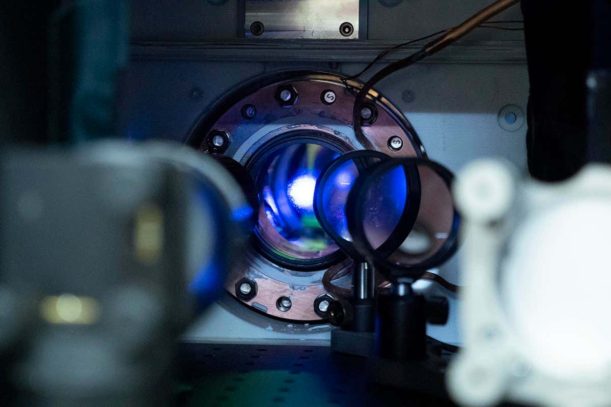 Atomic clock components sit in a laboratory setting, a blue light laser shines through a metal chamber, partially obscured by a pair of lenses