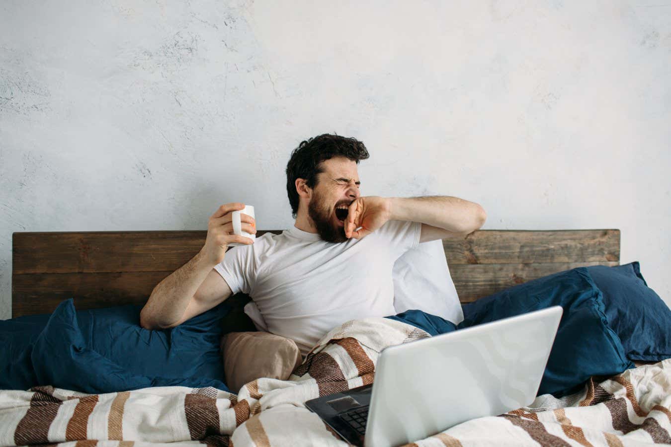 Handsome man lying in bed and warm blanket and stretching and yawning. Sleepy guy has laptop on the top and holding cup with hot tea or coffee in right hand; Shutterstock ID 622519436; purchase_order: -; job: -; client: -; other: -