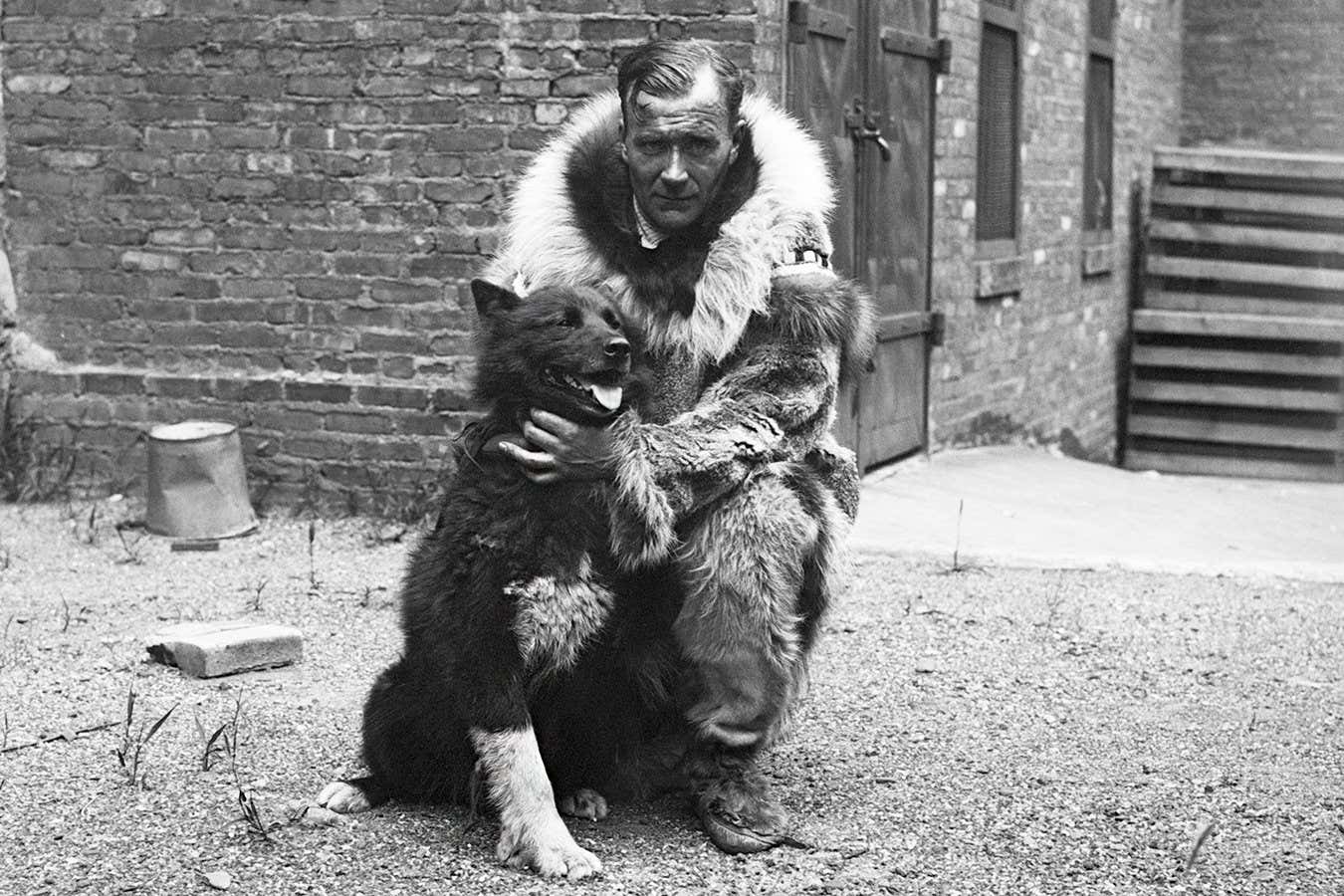 A black-and-white photo of a man dressed in warm furs crouching down next to Balto, a dog that appears to be a dark-furred husky type dog. Behind them is a brick building.