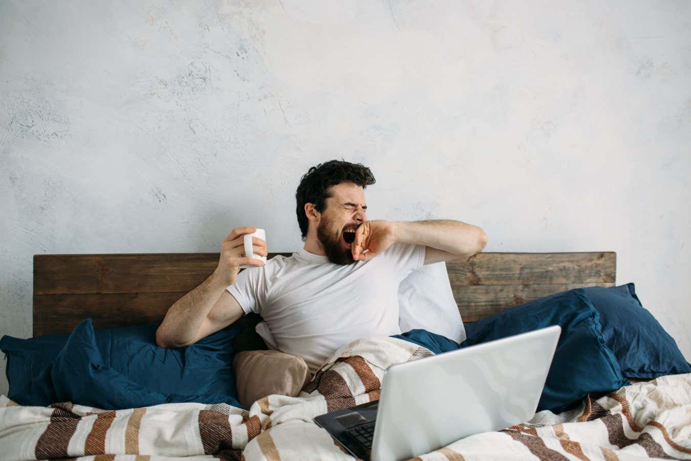 Handsome man lying in bed and warm blanket and stretching and yawning. Sleepy guy has laptop on the top and holding cup with hot tea or coffee in right hand; Shutterstock ID 622519436; purchase_order: -; job: -; client: -; other: -