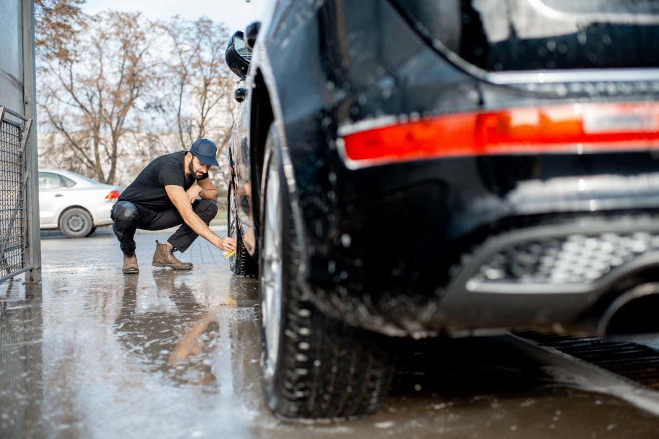 R2YAK1 Professional washer in black uniform and cap wiping with sponge car wheel of a luxury car during the washing process outdoors
