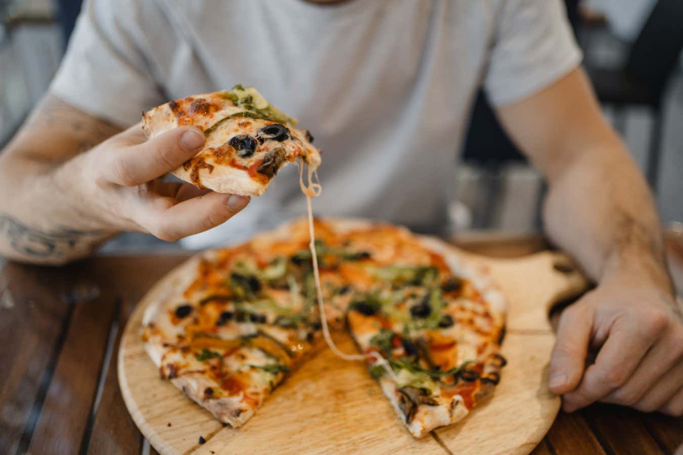 white young man with hat eating a pizza with vegetables on a wooden table
