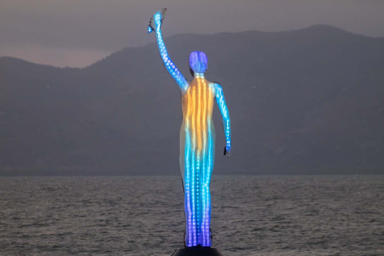 Ocean Siren, a coastal sculpture linked to a weather station on the Great Barrier Reef. As sea temperatures change the siren changes colour to reflect the data. Prolonged high temperatures indicate a threat of coral bleaching. This modern day lighthouse creates a connection to an environment over 70km away, underwater. She holds a Bayler shell which is a traditional method of communication and historically used to bail out water from boats. A warning against the impacts of climate change.