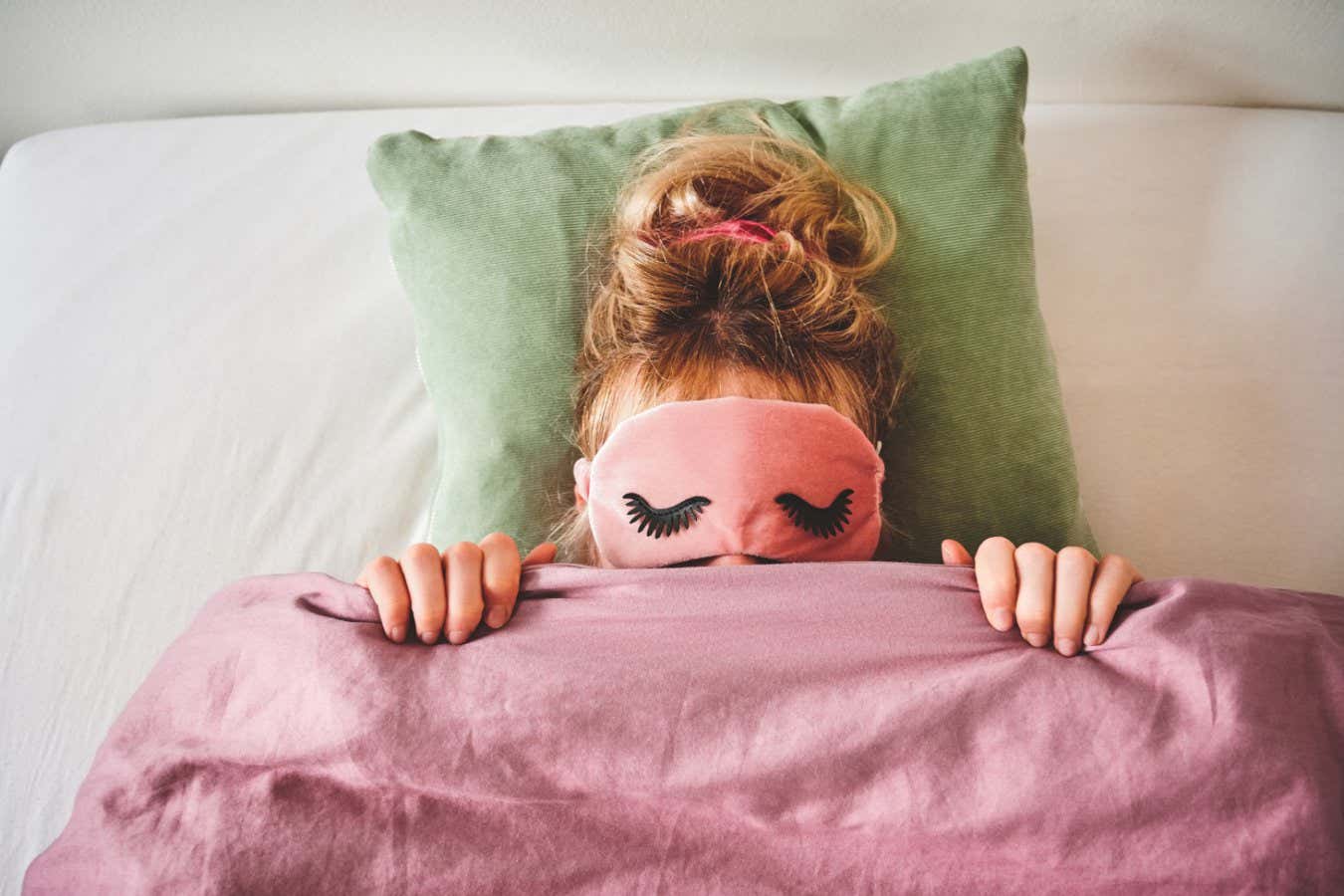 A woman in bed with a sleep mask on and covering her face with her duvet