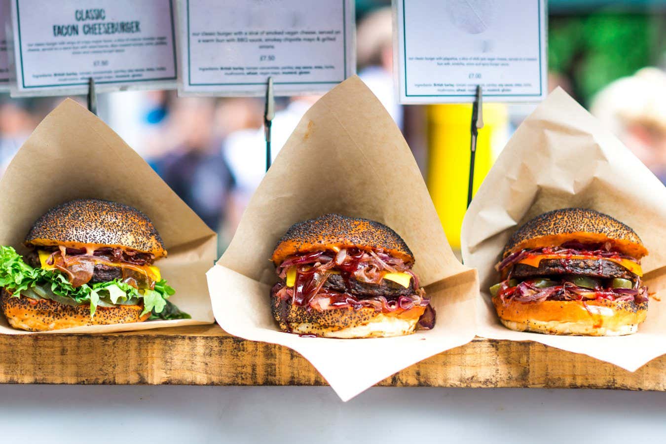 Close up image of a selection of freshly flame grilled burgers in a row on a wooden counter at a London street food market. Each of the burgers has its own label, on which is written the contents of the burger. The burgers are sandwiched between glazed buns, and presented on beds of fresh green lettuce and stuffed with melted cheese and red onion. Horizontal colour image with copy space and beautiful bokeh background.