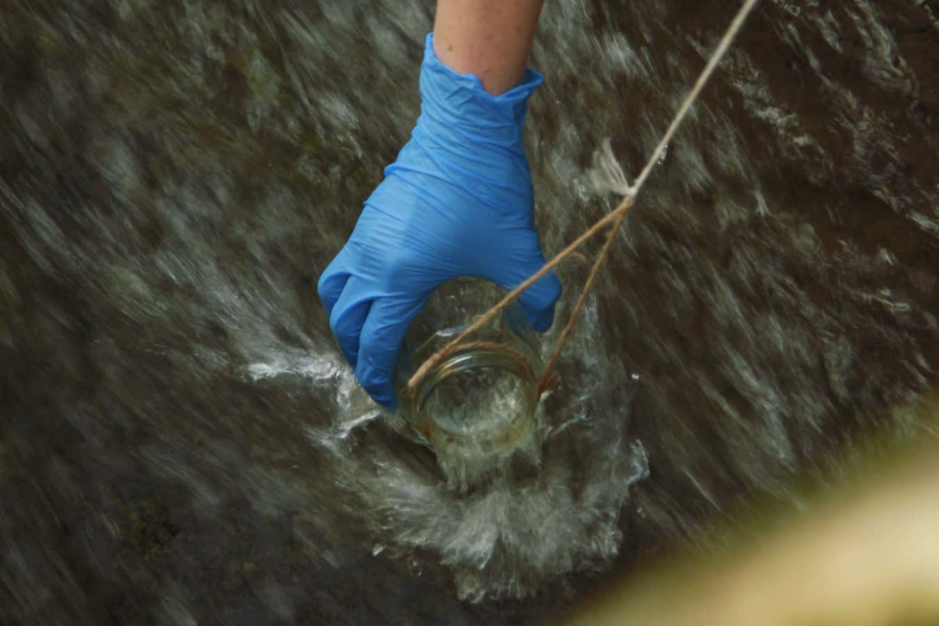 A blue gloved hand dips a glass container on a string into flowing water