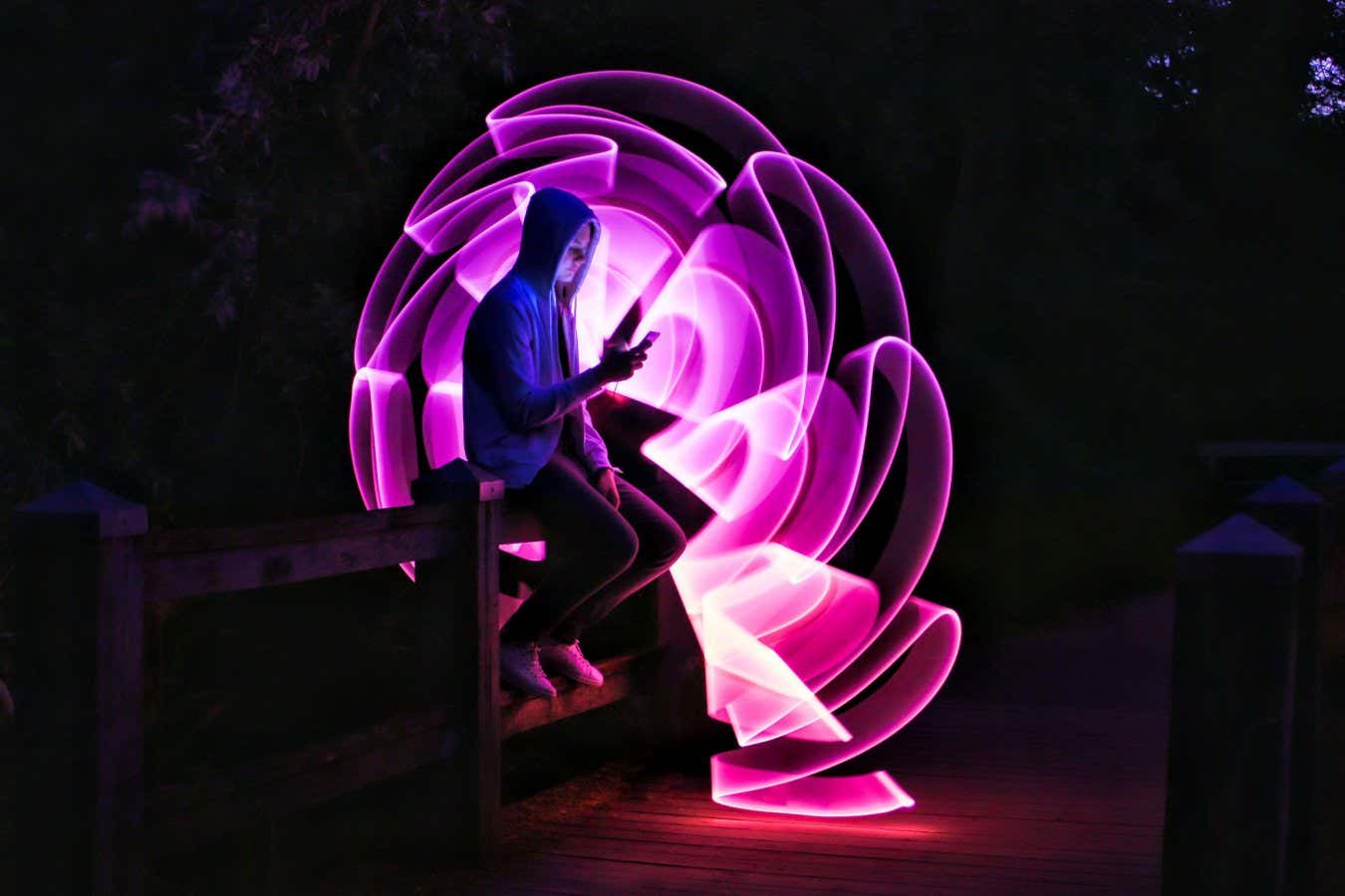 2C72973 Boy sitting on a bridge with his smartphone in his hand. Violet light effect with a light saber in lightpainting.