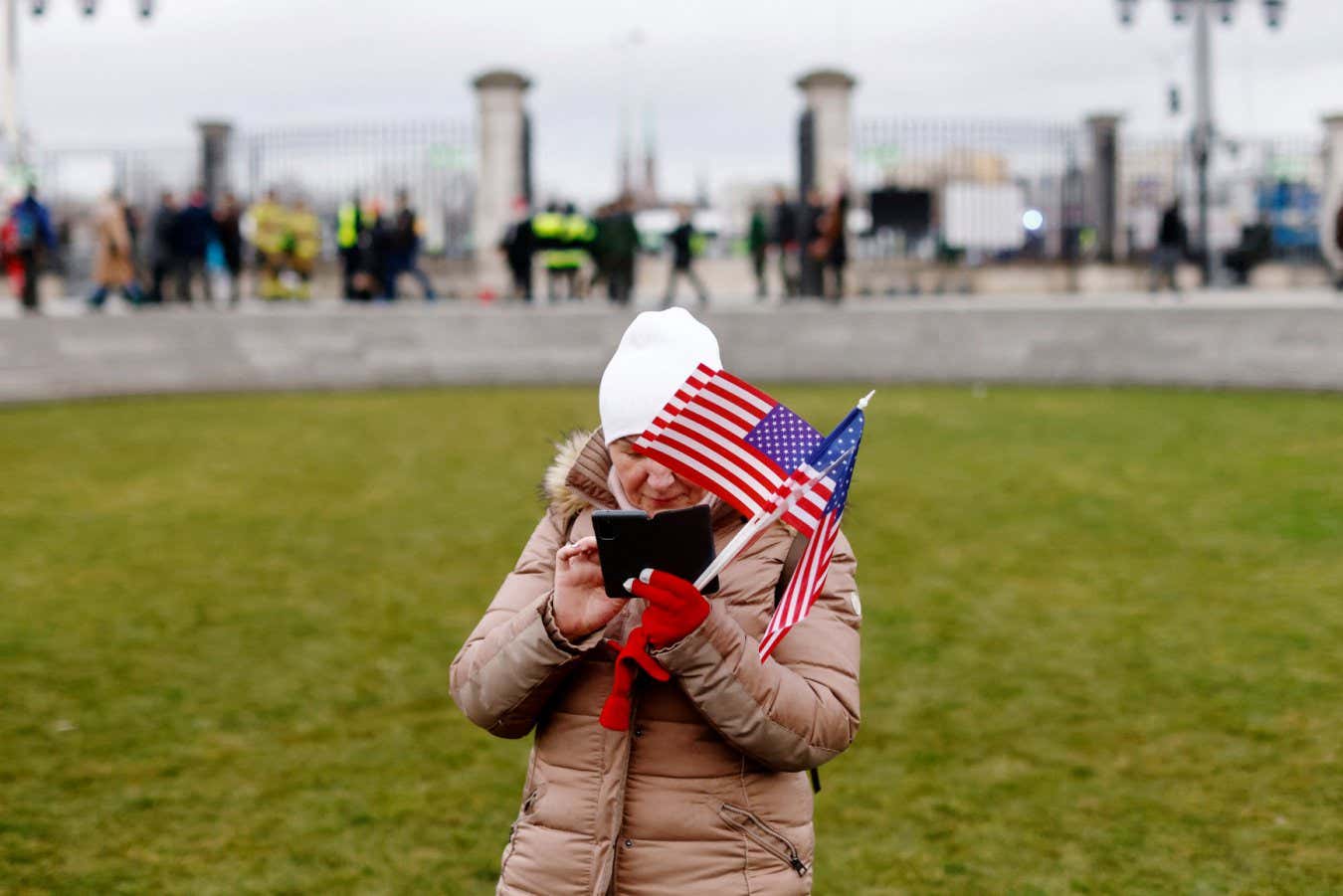 A woman holding several small flags of the United States checks her mobile phone while waiting with other onlookers prior to the arrival of the US President in Warsaw on February 21, 2023. - US President Biden is due to deliver a speech in Warsaw later on February 21 at Royal Warsaw Castle Gardens. (Photo by Wojtek Radwanski / AFP) (Photo by WOJTEK RADWANSKI/AFP via Getty Images)