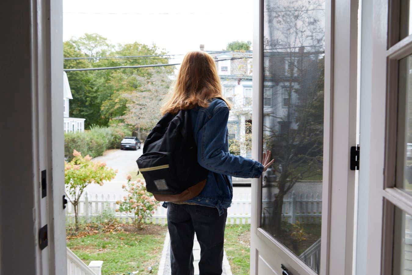 Teenage girl on her way to school going out her front door. We see her from behind going out the door and holding it open. She is wearing a back pack and a denim jacket. Shot from the knees up.
